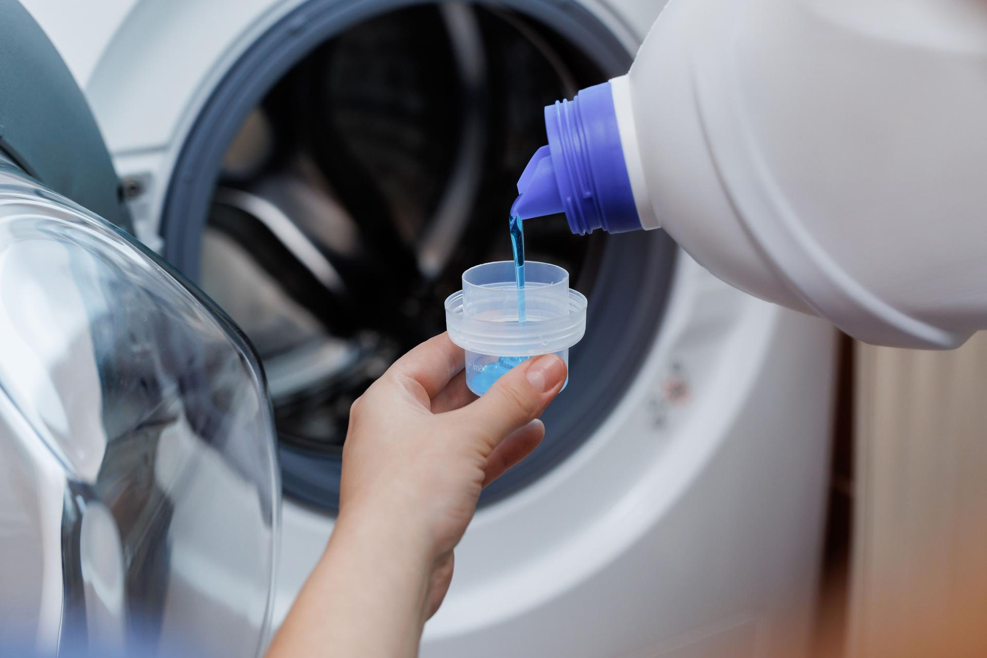 Close up shot of a woman hands squeezing liquid gel laundry detergent from a plastic bottle into a small container, with a washing machine in the background.