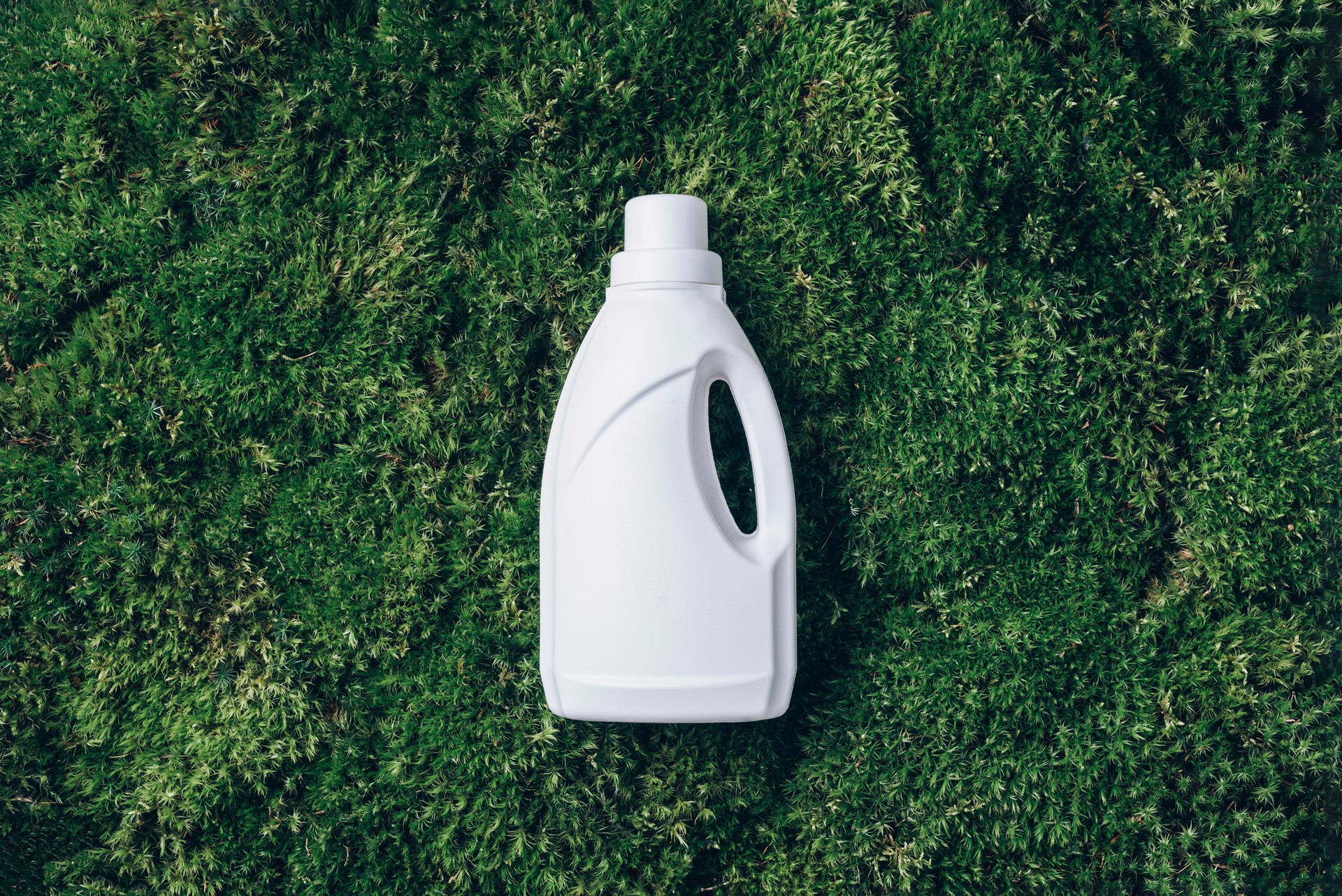 White plastic bottle of cleaning product, household chemicals or liquid laundry detergent on green grass, moss background. Top view. Flat lay. Copy space. Detergent bottle